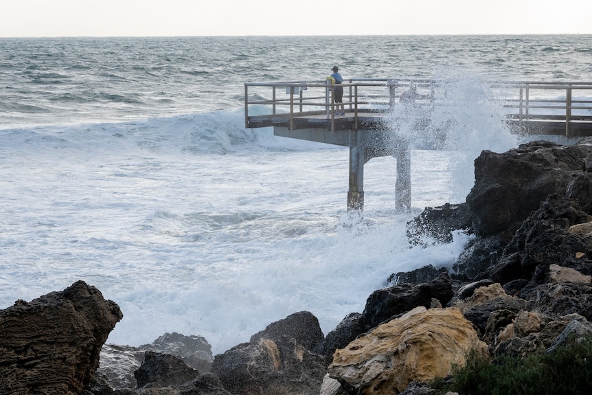 A lone child on a jetty that juts out into the ocean, with stormy weather and surf conditions, waves crashing against rocks.