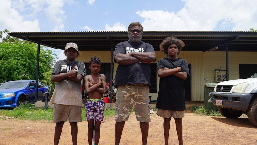 An Indigenous man standing with his family in front of a house on a sunny day.