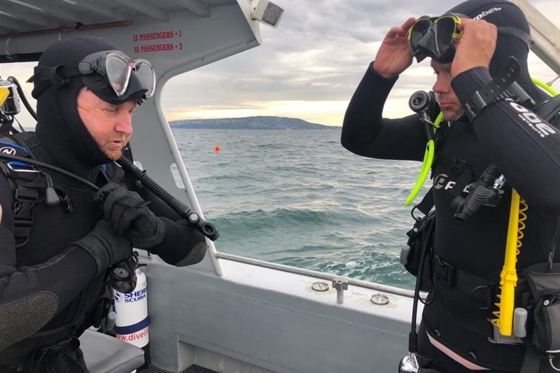 Two men in diving suits stand on a boat.