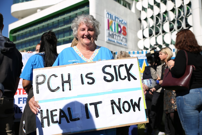 Jane holds a sign saying 'PCH is sick heal it now' while standing in front of the Perth Children's Hospital at a rally.