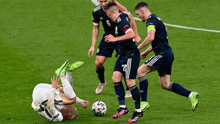 Three Scotland players stand over Phil Foden as he rolls on the ground.