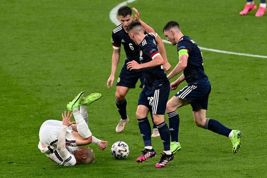 Three Scotland players stand over Phil Foden as he rolls on the ground.