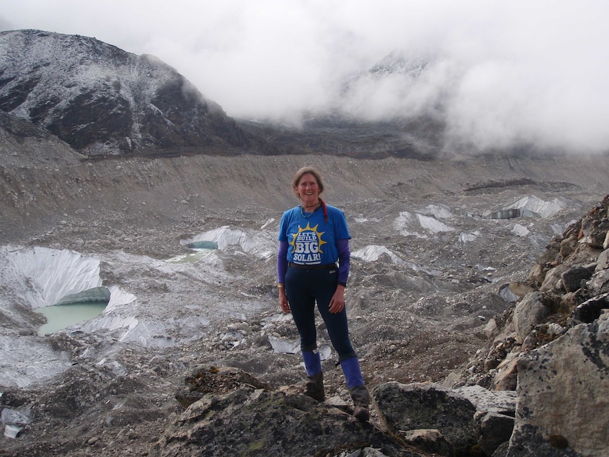 A woman stands on a grey rocky surface, with mist and ice in the background.