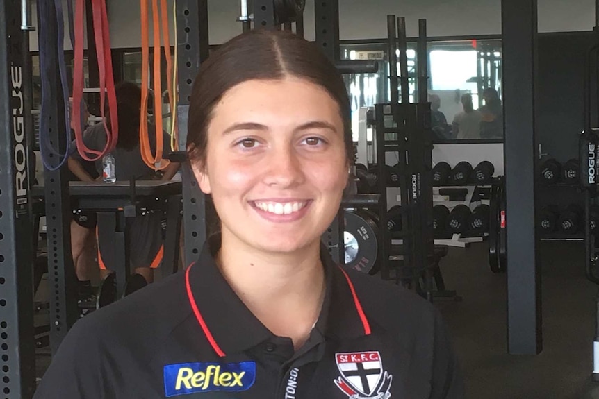 A young woman smiles at the camera in a gym with weights and equipment behind her.