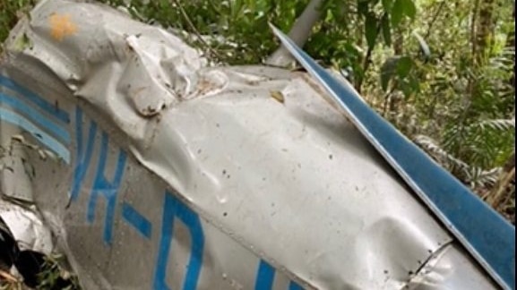 Plane wreckage is seen among trees and undergrowth. The plane was silver with blue writing that reads VHDJU and has stripes.