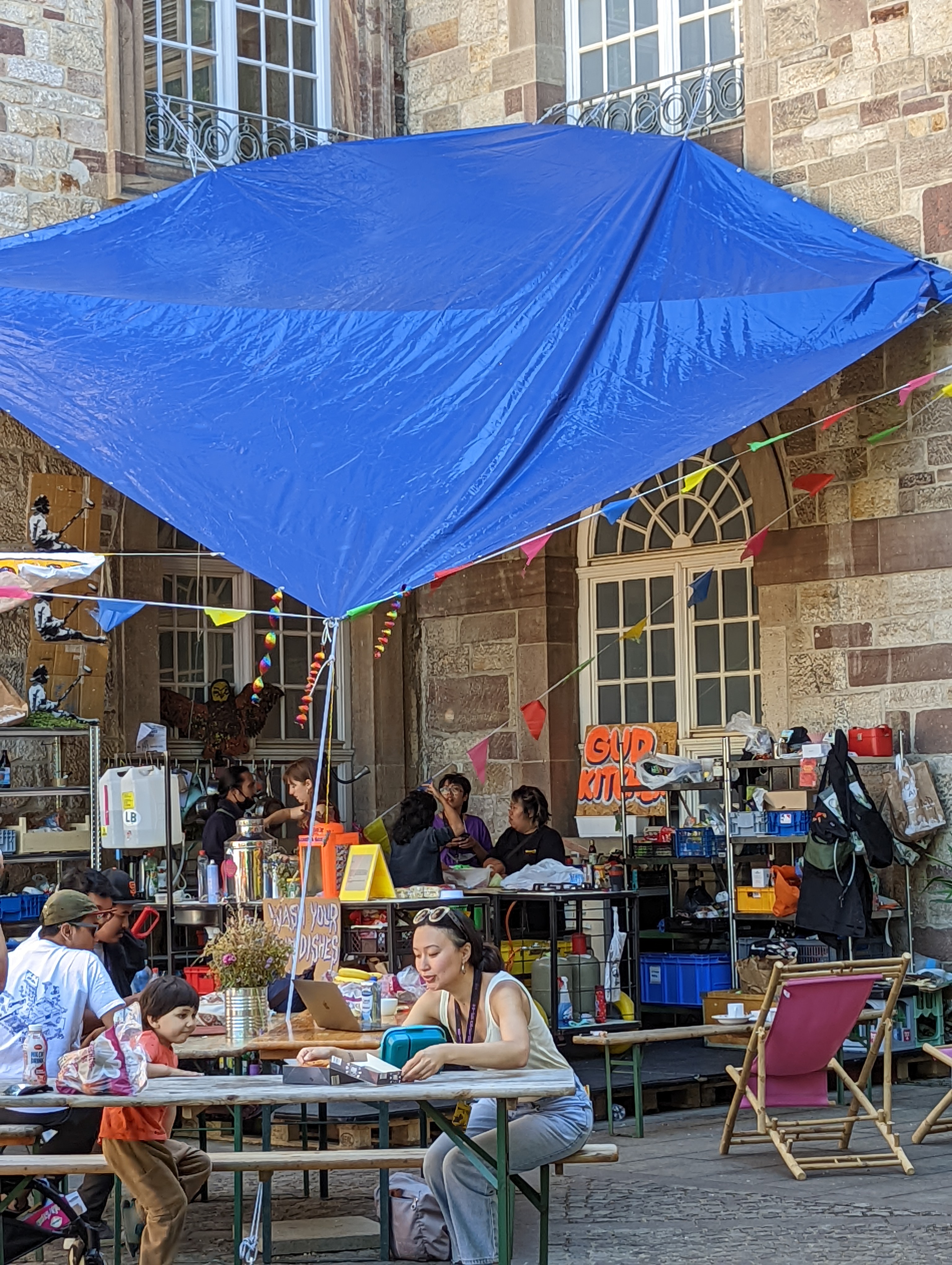 People sit on long benches at tables in a trinket-filled courtyard covered partially by a large blue tarp.