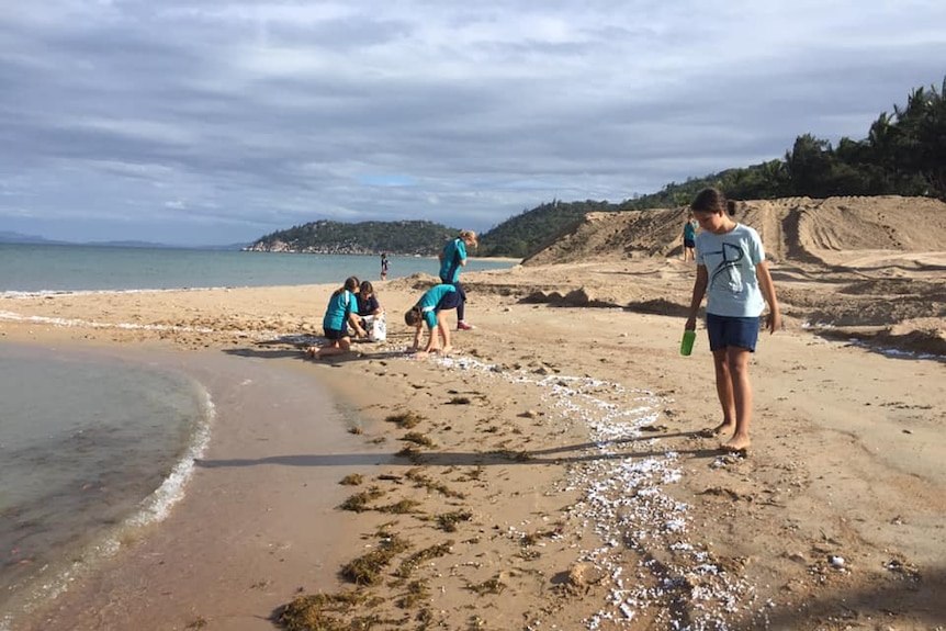 Five people, including kids in school uniform, bend down on the beach to clean up the polystyrene balls.