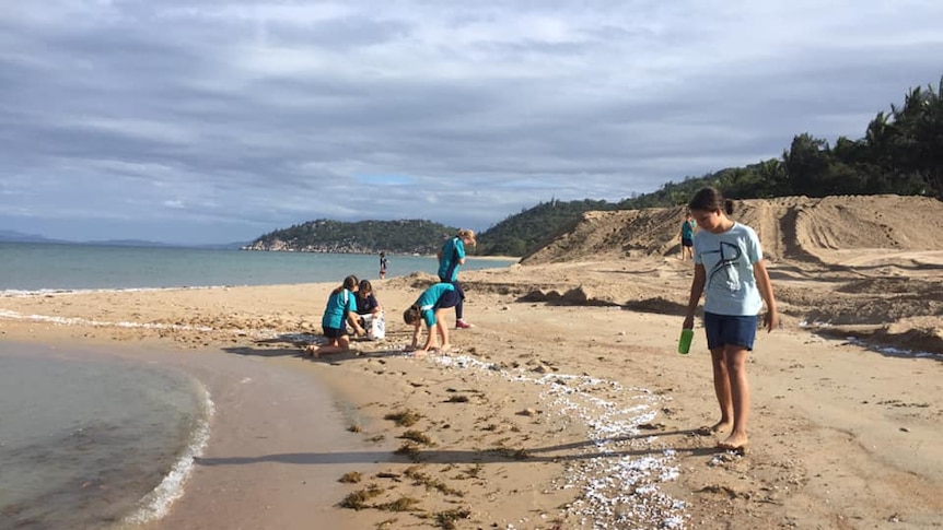 Five people, including kids in school uniform, bend down on the beach to clean up the polystyrene balls.