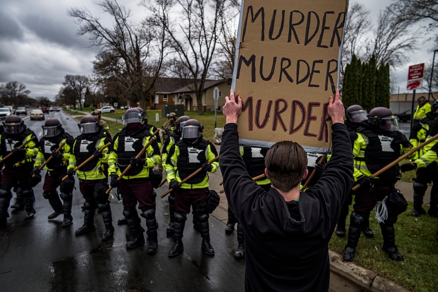 Protesters confront police wearing riot gear with a sign that says "murder, murder, murder".