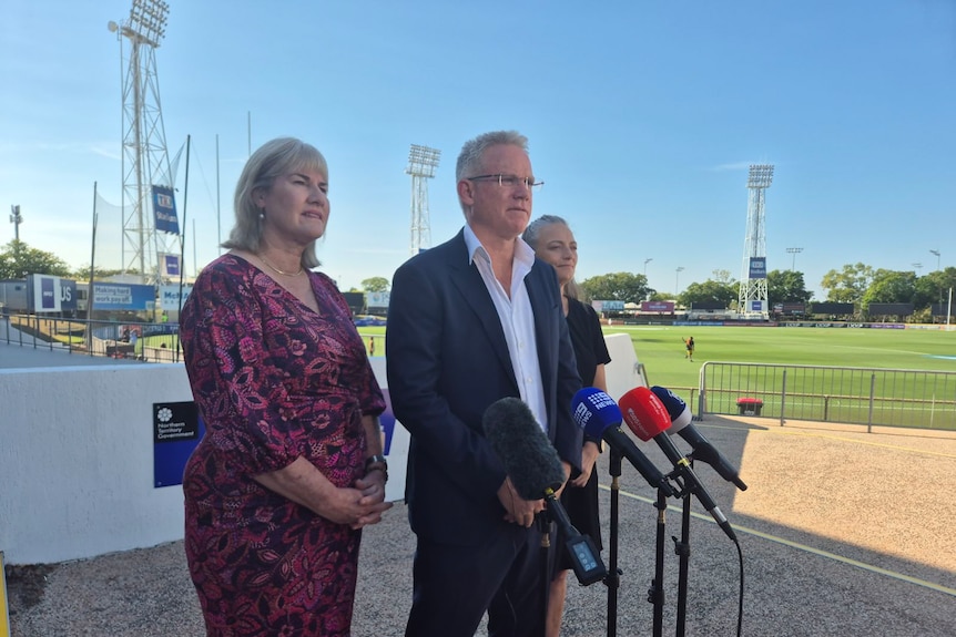 Three people stand in front of microphones at press conference next to football oval