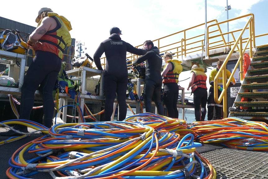 People in wetsuits working on a marine platform, lots of equipment lying around.