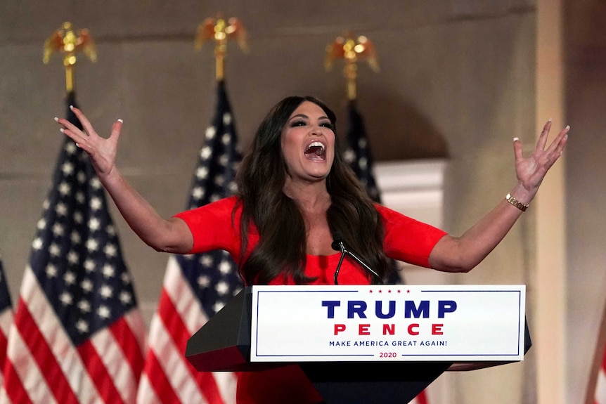 A woman with dark hair smiles with her mouth open and her hands in the air at a podium that read Trump Pence
