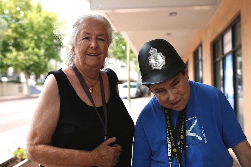 An older woman stands on a footpath next to her daughter who is wearing a blue shirt and toy police helmet.