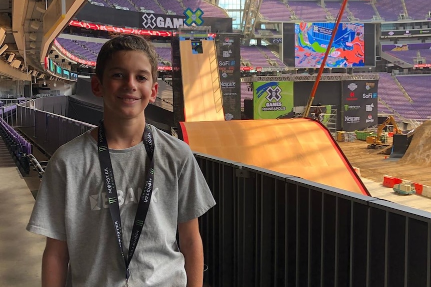 A smiling boy stands looking at the camera with X-Games signs and ramps in the background.