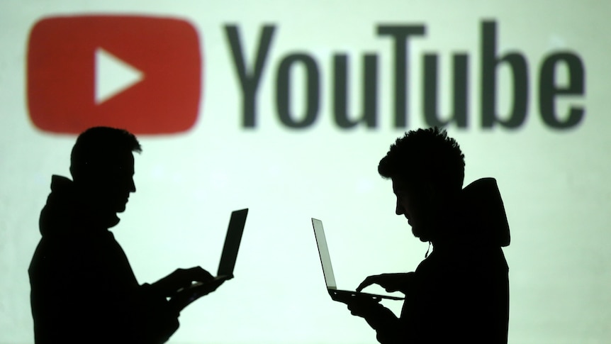 Silhouettes of mobile users are seen in front of a screen projection of the YouTube logo.
