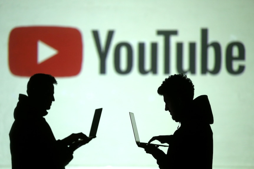 Silhouettes of mobile users are seen in front of a screen projection of the YouTube logo.