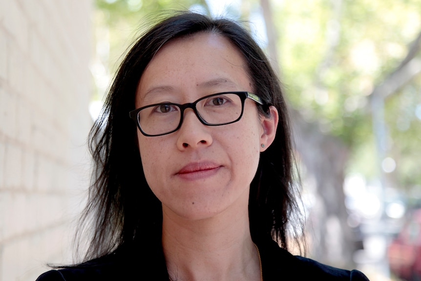 Colour photo of writer Rebecca Lim wearing striped shirt and black blazer standing outdoors next to white brick wall.