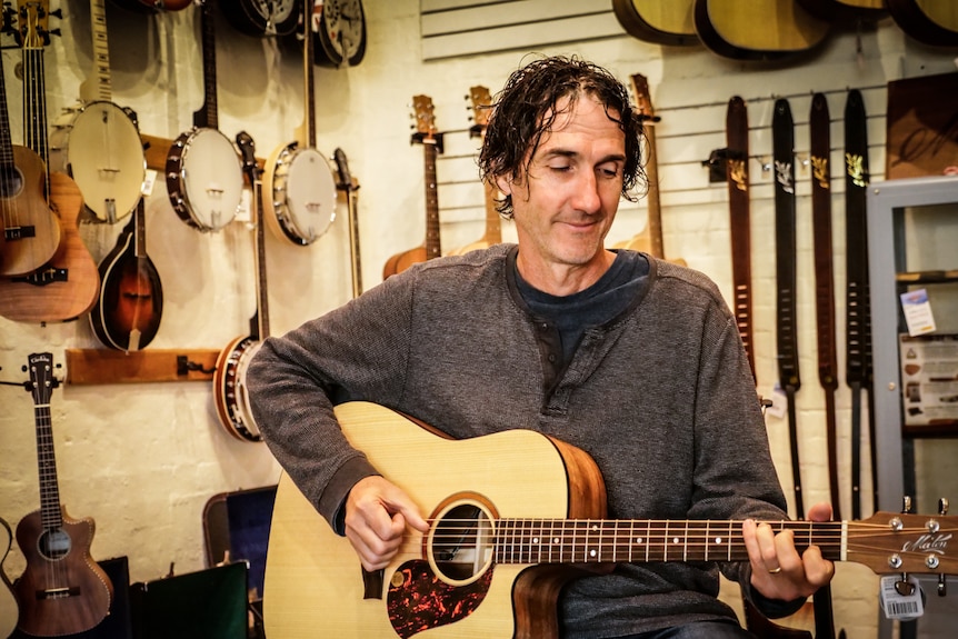 A man sits in a room full of guitars playing a guitar.