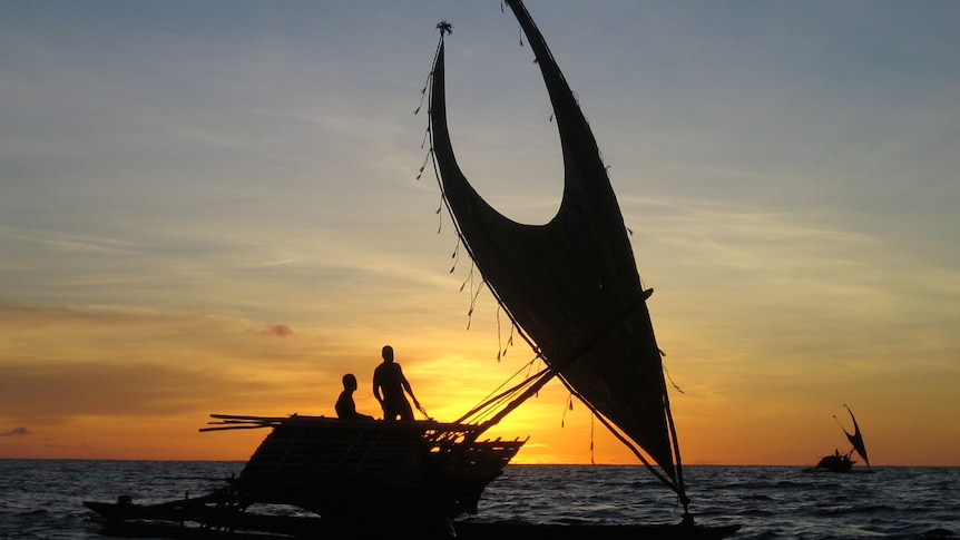 Two people stand on a traditional boat, silhouetted at sunset on the water.