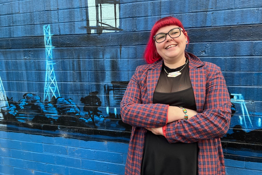 A woman with her arms folded standing in front of a mural depicting scenes from the local power industry