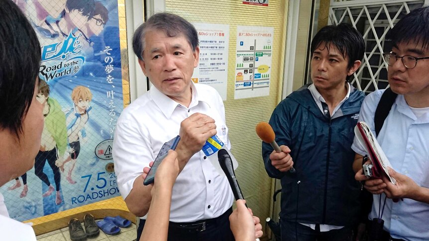 A man in a white shirt holds a drink bottle as he speaks to a throng of reporters inside an anime office.