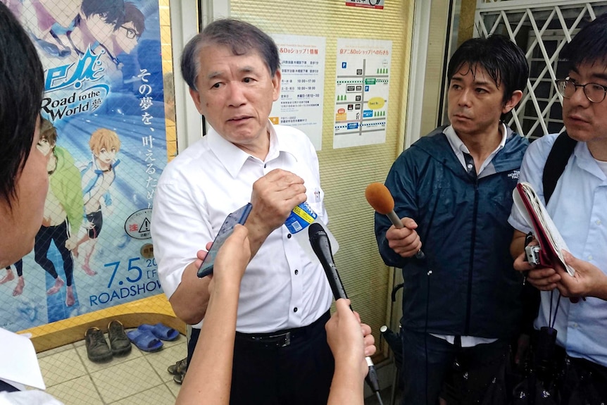A man in a white shirt holds a drink bottle as he speaks to a throng of reporters inside an anime office.