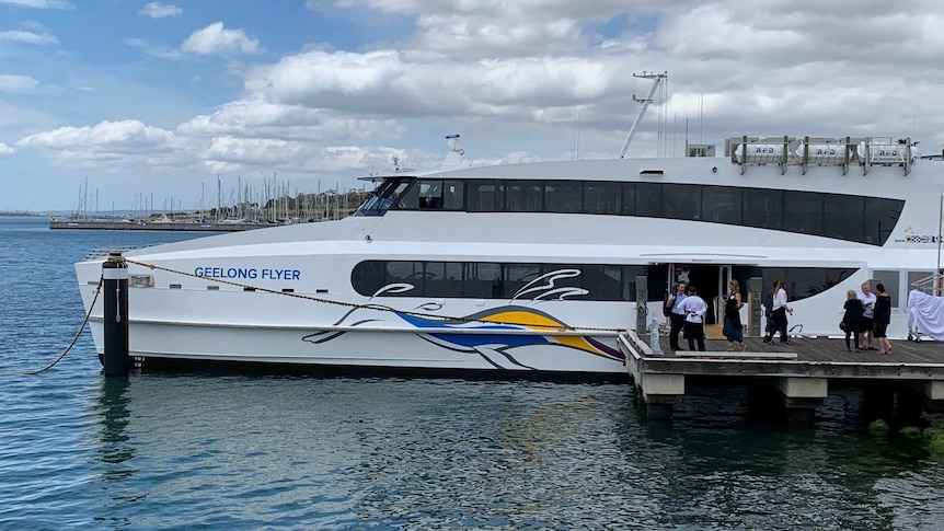 A modern two-level white ferry is docked by a pier. A yacht club is visible  in the background.