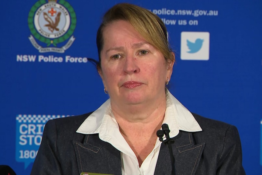 A solemn-looking middle-aged woman stands in front of a NSW Police-branded backdrop.