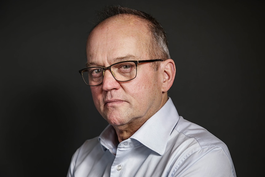 Colour close-up photo of Australia Council CEO Adrian Collette AM posing in front of grey background.