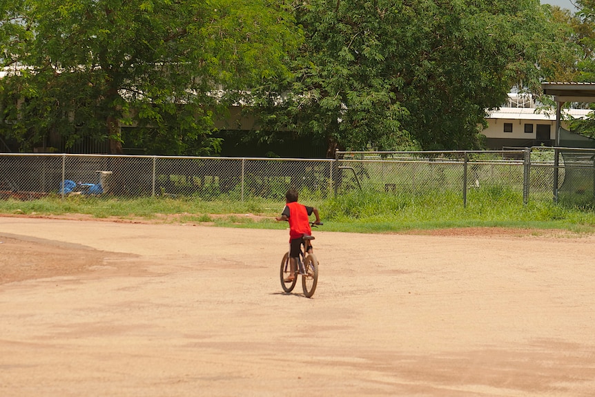 A kid in a bright red shirt riding a bike on a dirt road, trees in the background.