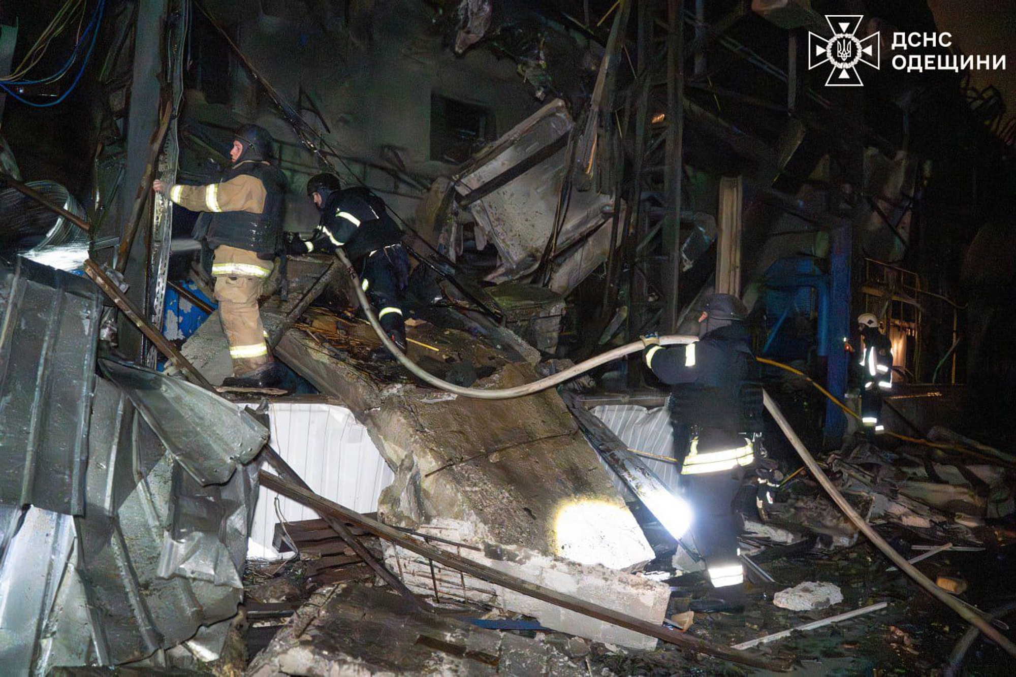 Los bomberos trepan por encima de los escombros de un edificio afectado por un ataque aéreo, llevando una manguera.