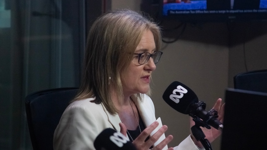 A woman with blonde hair, white blazer and black glasses gesticulates with her hands in front of a microphone.