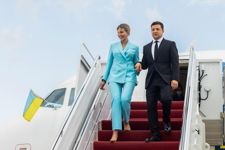 Volodymyr Zelenskyy and his wife Olena walk off a plan down some stairs holding hands. She is in a turquoise suit, he in black