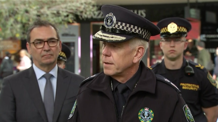 A man wearing a police hat with a man in a suit to the left and a man in a police officer's uniform with gold markings