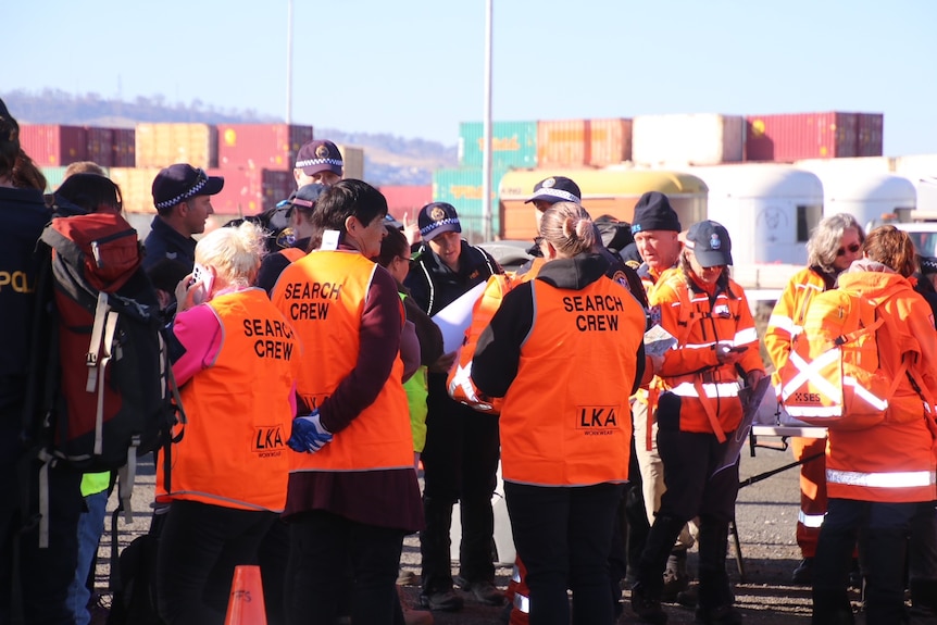 A group of people wearing orange Search Crew vests and jackets stand with police in front of shipping containers. 
