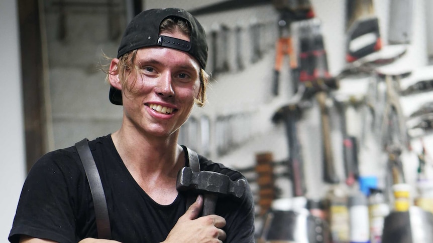 A young man sits in his armourer workshop smiling.