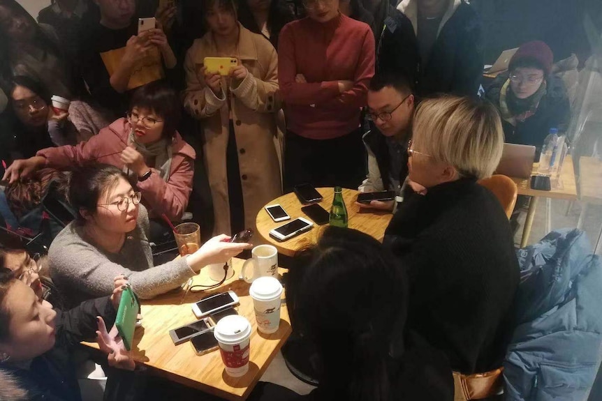 A group of people stand around a woman and take photographs with iphones while she sits and talks into a microphone