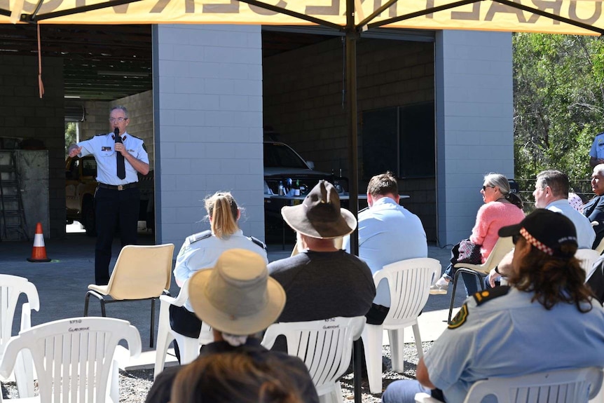 A man with grey hair and glasses stands in uniform addressing a seated crowd outside Eurimbula Rural Fire station