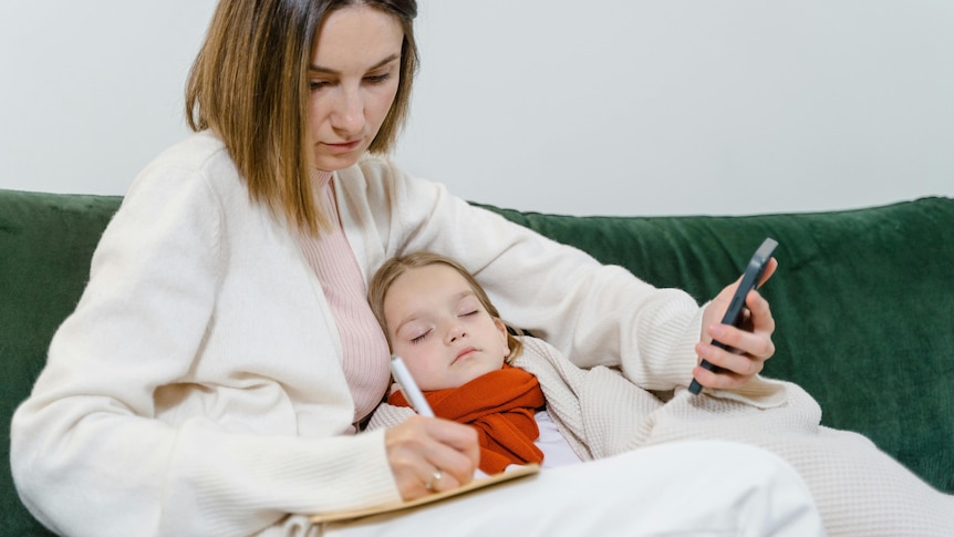 A mother caring for a sick child, holding a phone and writing down something.