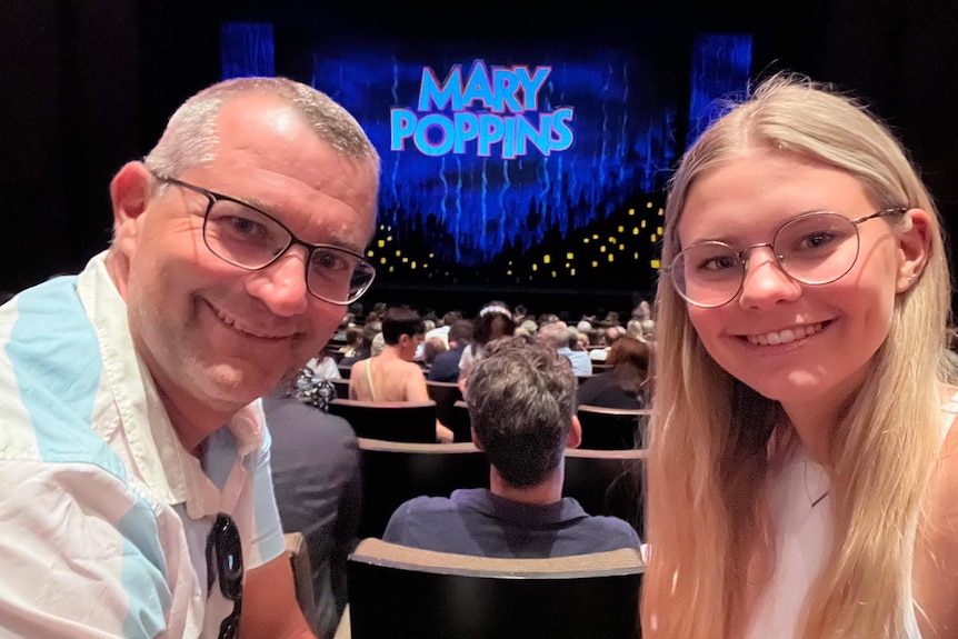 White, middle-aged man with long-haired blonde teenage daughter in front of Mary Poppins theatre curtain.