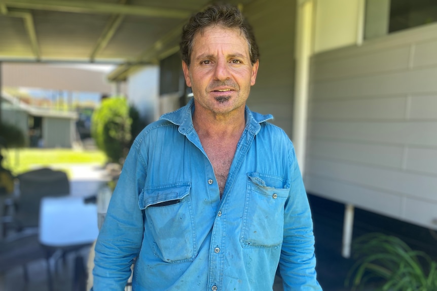 Joe Bugeja stands in front of a blurred background of the outside of a house, wearing a long-sleeved blue shirt