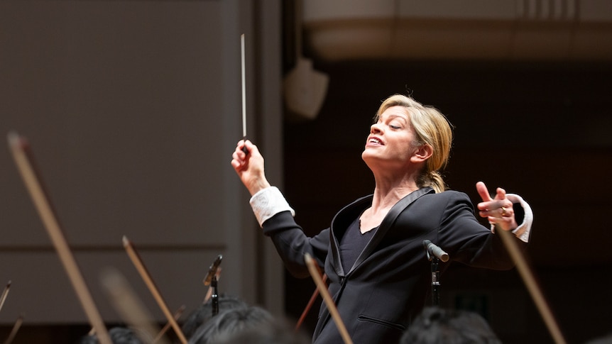 Maestro Keri-Lynn Wilson smiling widely holds her conductor's baton up high as she faces musicians before her.