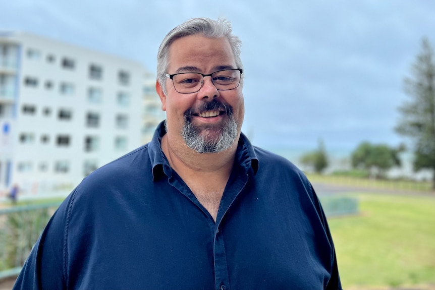 Hombre blanco con gafas y barba gris parado frente a un edificio frente al mar. 