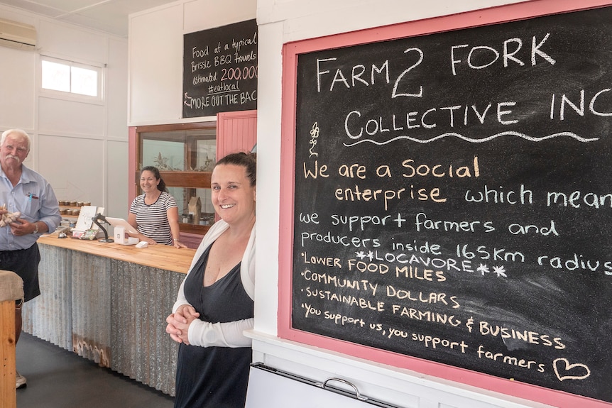 A man and two women stand in a shop next to a chalkboard filled with messages about eating local food.