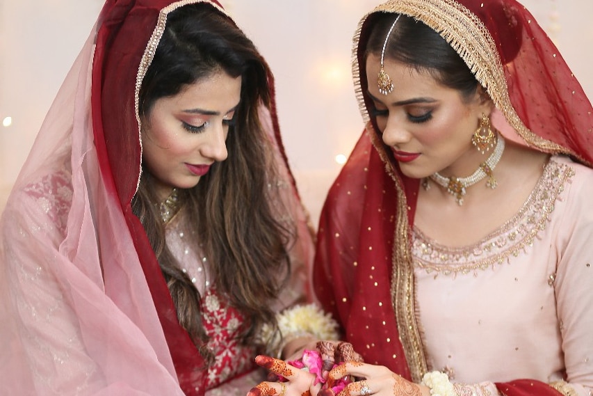 The bride, Hani Ali, seated with a bridesmaids, both dressed in traditional Pakistani wedding attire.