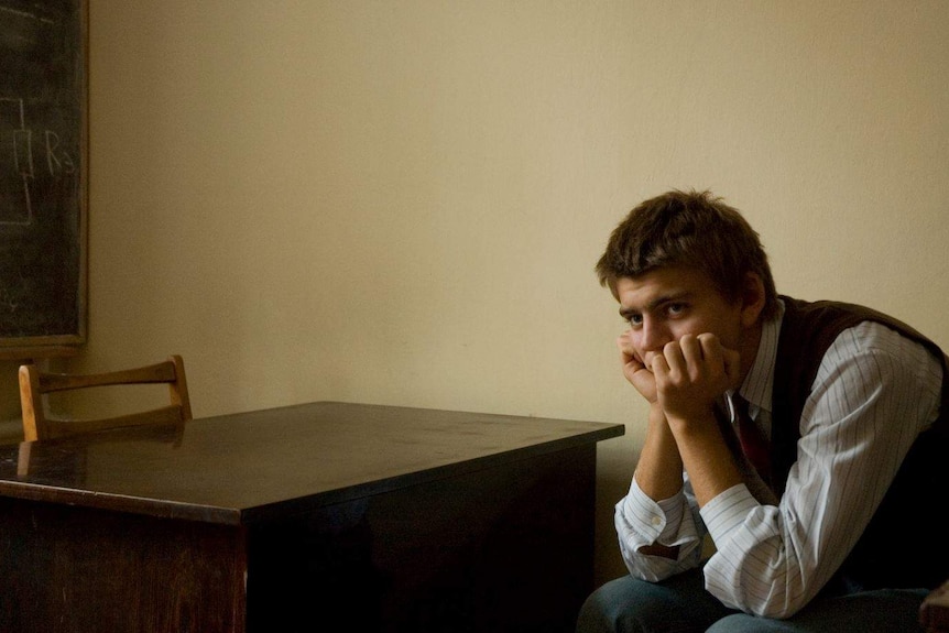 A young man sits in an empty room at a wooden desk, with his hands covering his mouth staring into the distance.