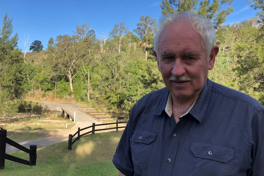 A man with white hair wearing a blue work shirt standing in a park.