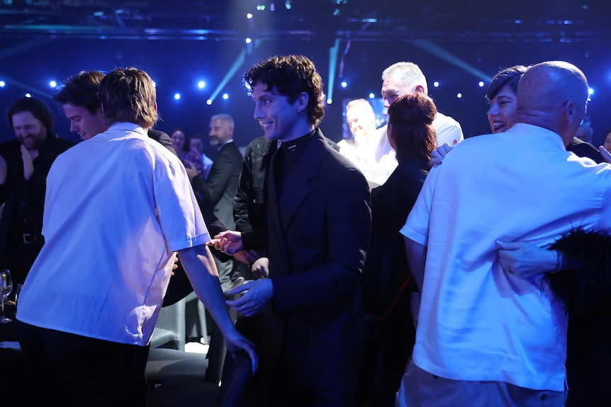 Troye Sivan smiles as he stands from his seat as people around him celebrate at an awards night