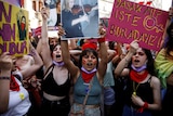 Three women, at the front of a crowd of many, scream and hold up colourful signs and photos of women.