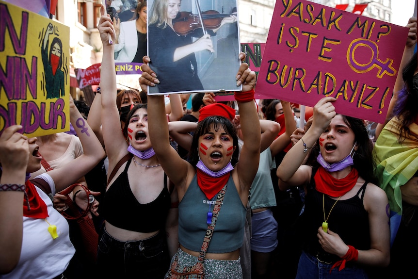 Three women, at the front of a crowd of many, scream and hold up colourful signs and photos of women.